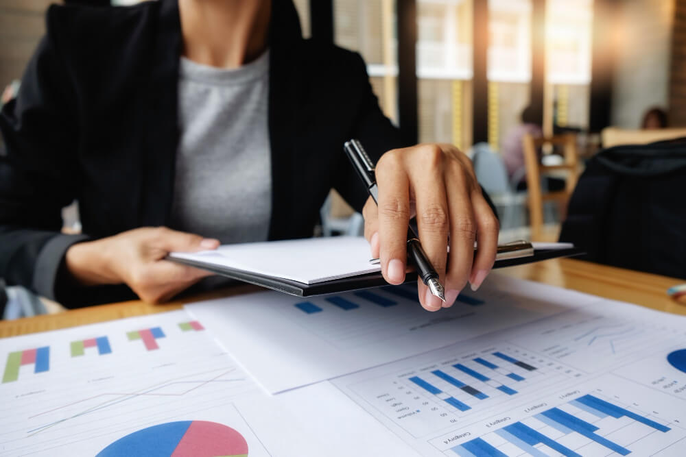 Mão segurando caneta sobre planilhas com indicadores financeiros, ilustrando a análise de um relatório de vendas em ambiente corporativo.