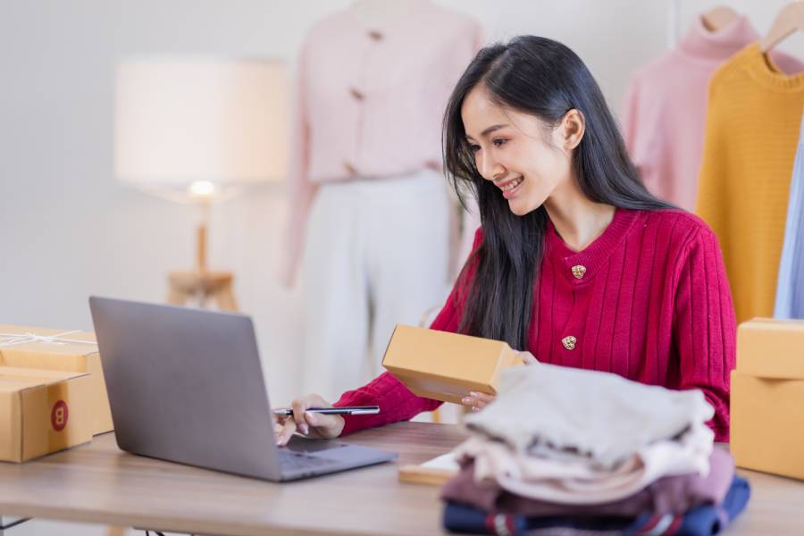 Mulher sorrindo gerencia gestão de catálogo em marketplace com laptop, caixas de envio e roupas ao fundo. 