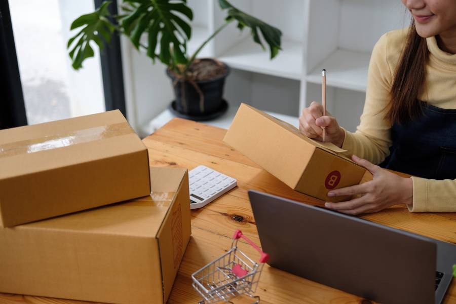 Pessoa anotando informações em caixa de papelão com laptop aberto 
